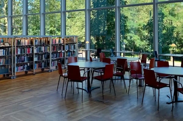 Une grande salle de bibliothèque avec de hautes fenêtres offrant une vue sur un paysage verdoyant. La salle est équipée de plusieurs tables et chaises rouges, et des étagères remplies de livres sont disposées le long d'un mur. L'environnement lumineux et ouvert illustre le concept de bibliothèque comme troisième lieu, un espace accueillant pour la lecture, l'étude et la détente.