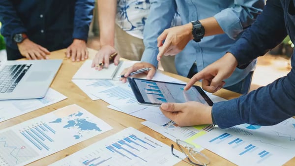 Un groupe de personnes examine des graphiques et des rapports sur une tablette et des documents papier sur une table. Un ordinateur portable est également présent. Cette image illustre une discussion sur les critères de positionnement dans le référencement Google en 2024.