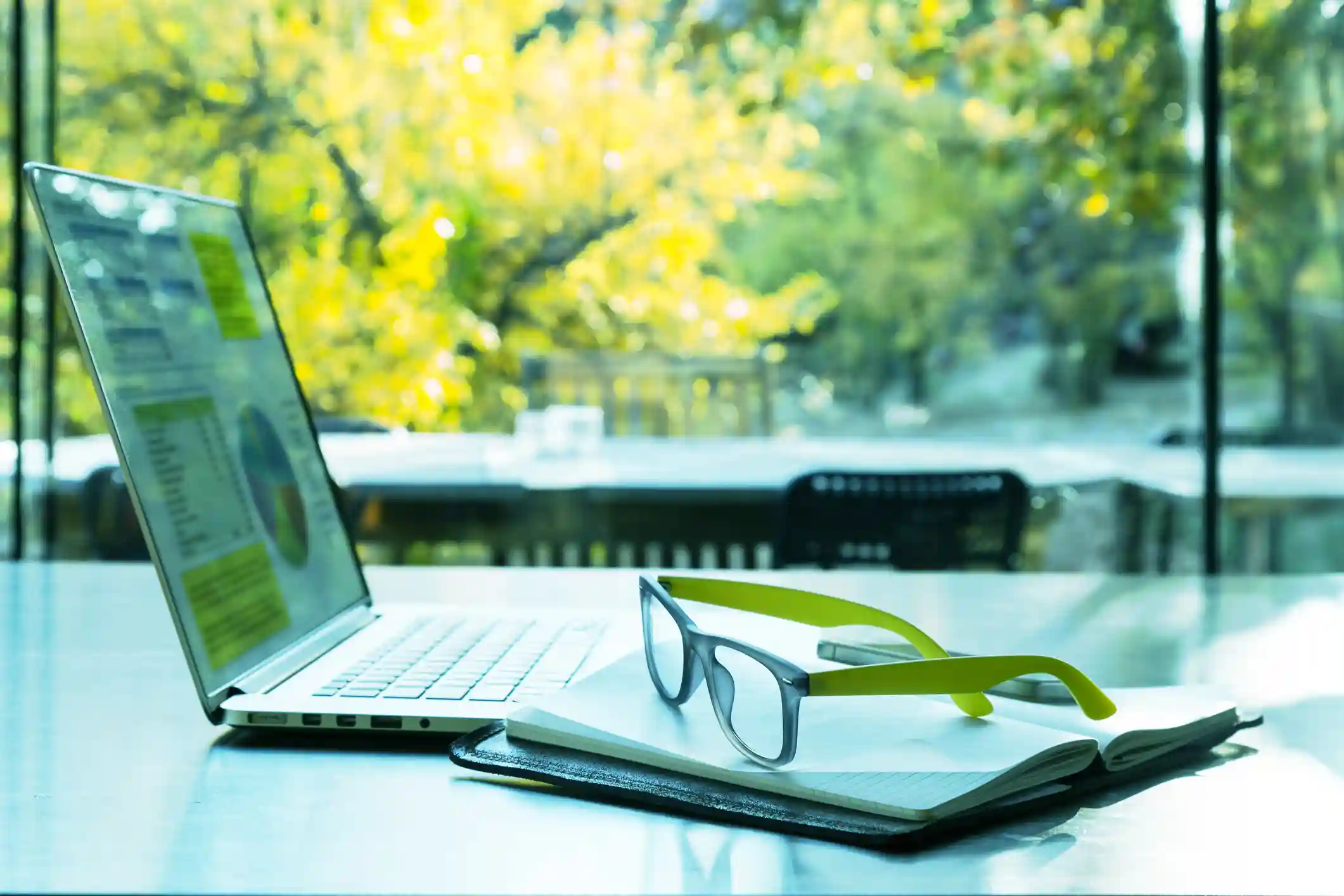 Un ordinateur portable ouvert affichant des graphiques, posé sur une table avec un carnet ouvert et une paire de lunettes. La scène est éclairée par la lumière naturelle provenant de grandes fenêtres donnant sur un paysage verdoyant, suggérant un environnement de travail axé sur l'éco-conception.