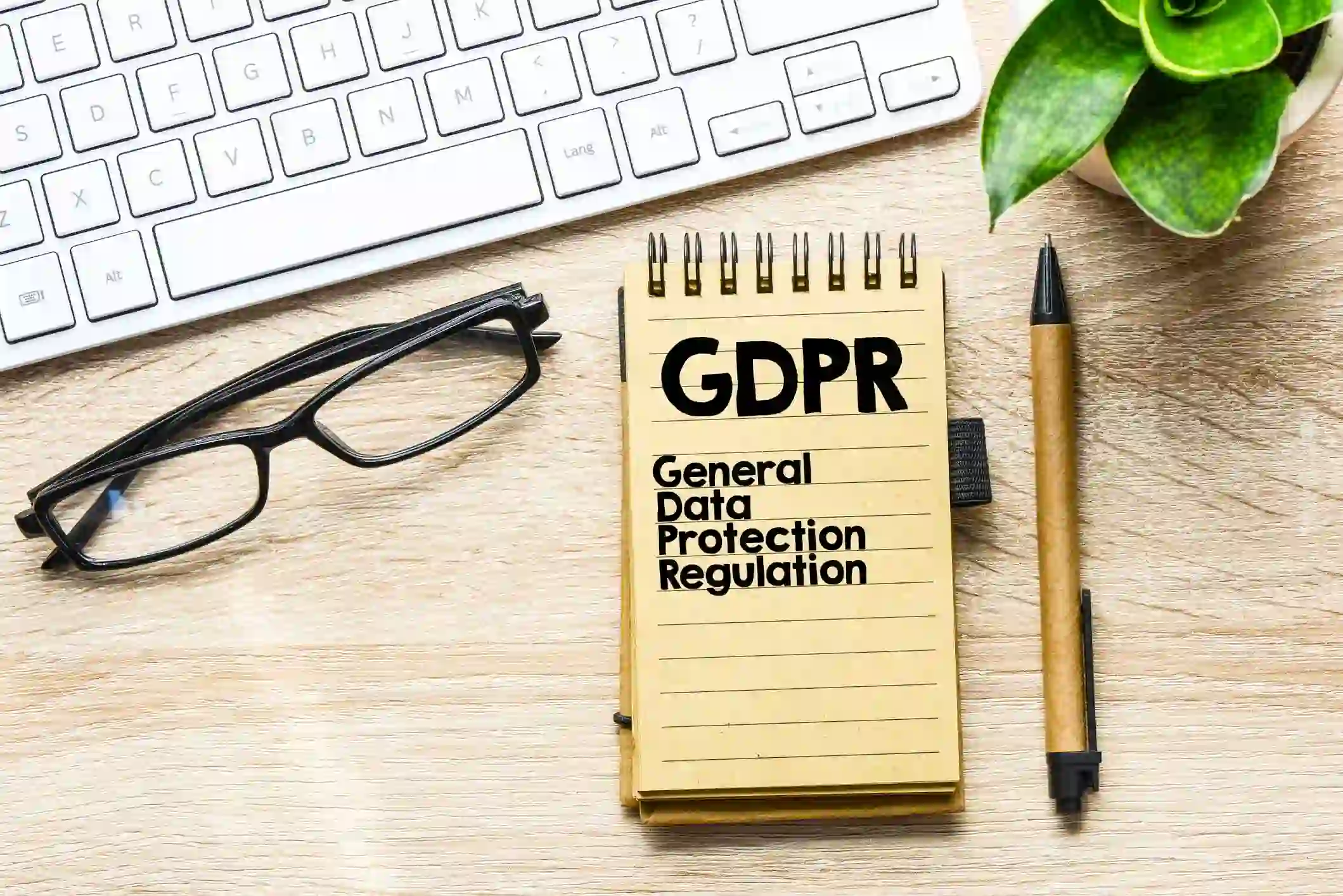Un carnet avec l'inscription 'GDPR General Data Protection Regulation' est posé sur un bureau en bois, à côté d'un clavier, d'une paire de lunettes et d'un stylo. Une plante verte est visible en arrière-plan, suggérant un environnement de travail axé sur la conformité au RGPD.