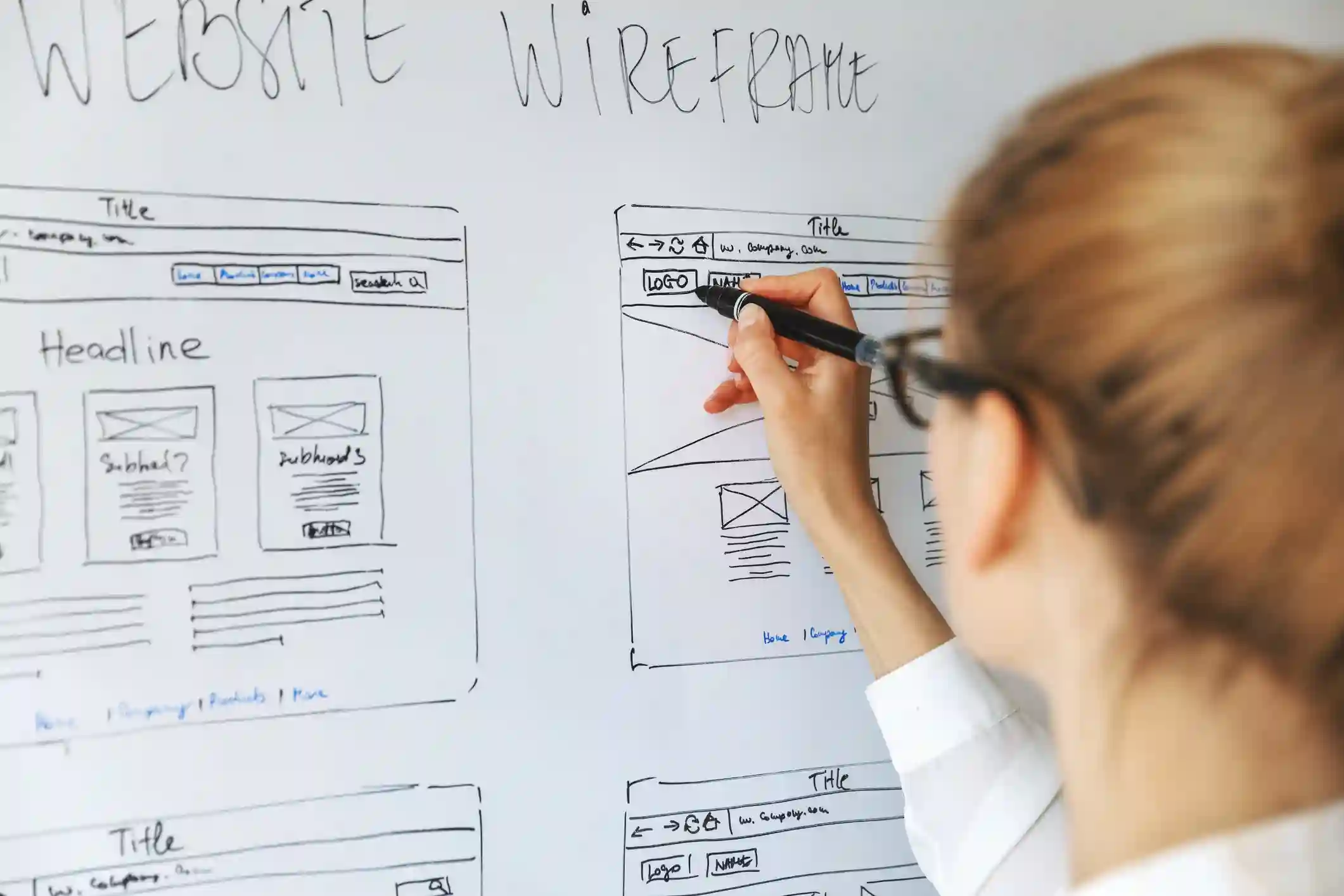 Une personne dessine des maquettes de sites web sur un tableau blanc, planifiant la structure et le contenu des pages. Le mot 'WEBSITE WIREFRAME' est écrit en haut du tableau, indiquant le processus de conception de l'expérience utilisateur (UX Writing).